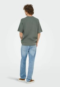 Grøn strikket kortærmet skjorte med rund hals, parret med lyseblå jeans og brune sko. Bagfra med synlige tatoveringer.