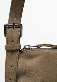 Borsa a tracolla in suede beige con un cinturino in metallo argentato, tracolla regolabile e chiusura con zip. Caratterizzata da una texture liscia e un design semplice.