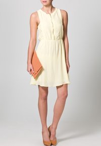 Un vestido sin mangas de color amarillo claro con cierre de botones en el frente, cintura entallada y falda fluida; combinado con un bolso naranja y tacones de mimbre en color marrón claro.