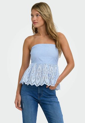 Jeune femme portant un haut rayé bleu et blanc sans bretelles avec un ourlet en dentelle ajourée, associé à un jean bleu, posant avec une main dans la poche.