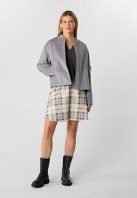 Giacca bomber grigia con zip frontale, sopra una maglia scura e una gonna a quadri crema in beige e grigio. Stivaletti neri alla caviglia con suole spesse.