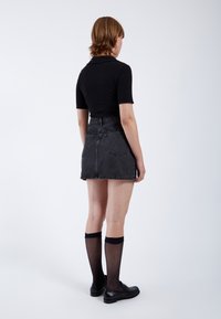 Zwarte geribbelde top met korte mouwen, hoge zwarte spijkerrok met rafelige zoom, gedragen met zwarte kniehoge sokken en loafers, gezien van achteren.