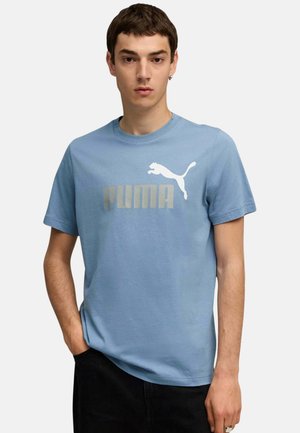 Joven con cabello corto y oscuro que lleva una camiseta azul claro de Puma y pantalones negros, de pie con una mano en el bolsillo frente a un fondo liso.