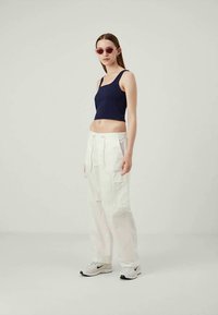 Young woman wearing pink cat-eye sunglasses, navy crop top, white cargo pants, and white Nike sneakers standing against plain background.