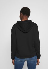 Il hoodie nero presenta una texture morbida, cappuccio con cordino regolabile e polsini a costina. Abbinato a jeans blu, con dettaglio della cucitura posteriore visibile.