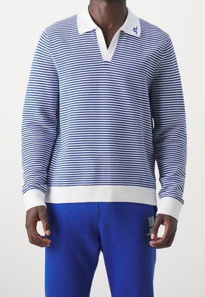 Hombre con polo de rayas azules y blancas de manga larga, con puños y cuello blancos, combinado con pantalones azules.