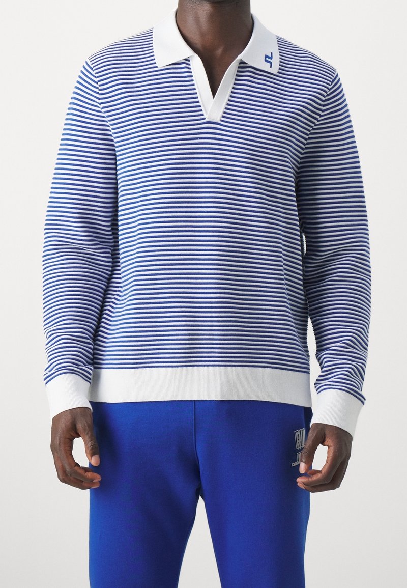 Homme portant un polo rayé bleu et blanc à manches longues avec des poignets et un col blancs, assorti à un pantalon bleu.