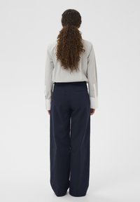 Femme aux longs cheveux bouclés attachés en queue de cheval, portant une chemise gris clair et un pantalon large bleu marine, debout de dos sur un fond blanc.