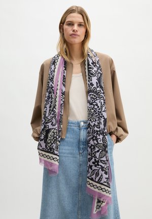Femme blonde portant une veste bomber beige, un haut blanc, une jupe en denim bleu et une longue écharpe noire à motifs floraux lilas. Mains dans les poches.
