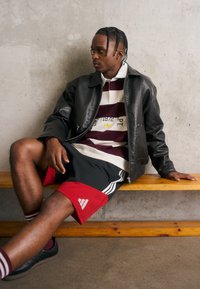 Μαύρο δερμάτινο μπουφάν πάνω από μαραoon και λευκή ριγέ πόλο μπλούζα; γκρίζα σορτς με κόκκινες λεπτομέρειες και πλευρικές ρίγες; καθισμένος σε μια ξύλινη παγκάκι.