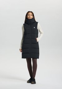 Gilet en duvet noir avec col montant, fermeture éclair sur le devant et poches latérales. Porté par-dessus un haut à manches longues léger et associé à un legging marron et des baskets.