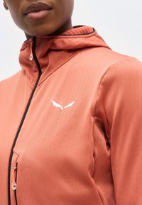 Κοντινή λήψη ενός ατόμου που φοράει ροζ-σολομό μπουφάν με φερμουάρ και κουκούλα, με λευκό λογότυπο σε σχήμα φτερού στο στήθος.