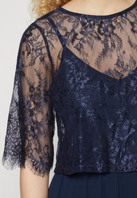 Λουλούδινη δαντελένια μπλούζα ναυτικού μπλε με διάφανες μανσέτες μήκους μέχρι τους αγκώνες και φράχτη στο τελείωμα, επενδυμένη από ένα συμπαγές σκούρο μπλε υπόστρωμα. Υφασμένη και λεπτή σχεδίαση.