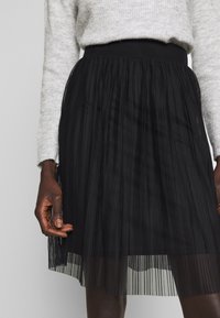 Jupe plissée noire avec une superposition de tulle texturé, dotée d'une taille élastique lisse. L'ourlet est transparent, révélant les couches en dessous.
