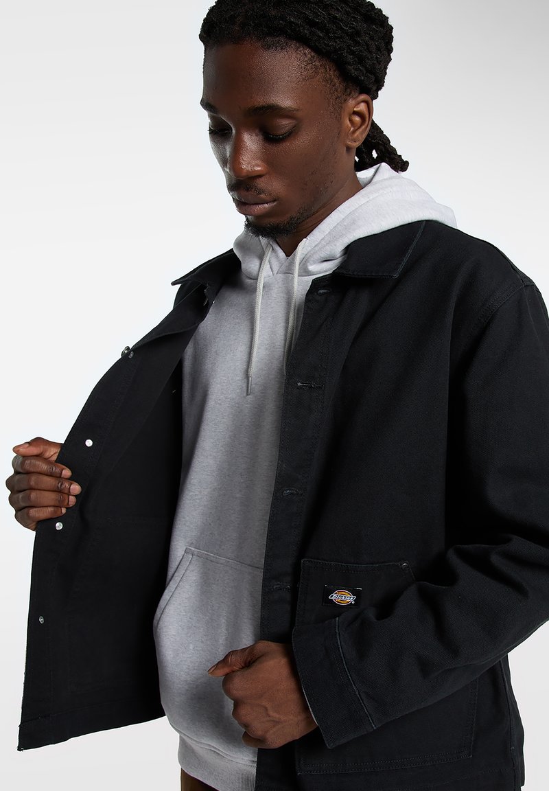 Giacca nera con bottoni sopra una felpa con cappuccio grigia. La giacca presenta un tessuto strutturato, tasche laterali e una patch con logo sulla tasca inferiore.