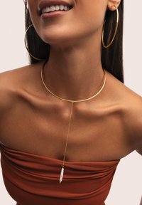 Goldene Choker-Halskette mit einer dünnen Kette und einem barocken Perlenanhänger, kombiniert mit großen goldenen Creolen, getragen über einem braunen Off-Shoulder-Oberteil.