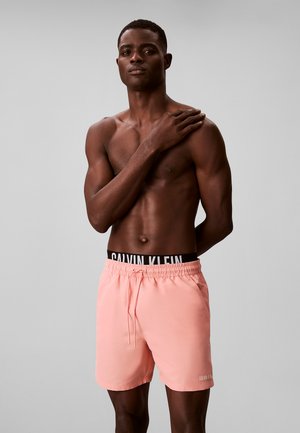 Muskuløs mand står bar overkrop med hånden på skulderen, iført lyserøde Calvin Klein badeshorts og sort trusse med taljebånd mod grå baggrund.