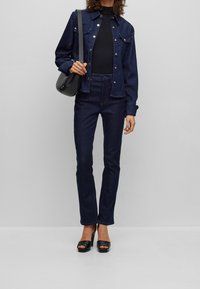 Giacca in denim scuro con bottoni dorati, abbinata a jeans scuri a vita alta. Top nero e tacchi aperti neri completano l'outfit. Inclusa borsa a tracolla.