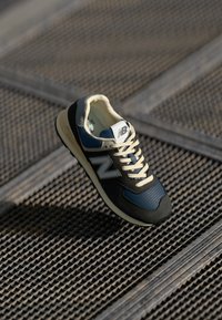 Zapatillas negras y marinas con un cuerpo de malla, acentos de ante y cordones crema. Presenta un destacado logo 'N' gris en el lateral. Punta redondeada.