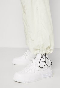 Vita lädersneakers med perforerat tårum, platta snören och texturerad gummisula, i kombination med ljusgräddafärgade smala byxor.