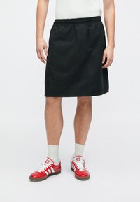 Svart, kne-lengde skjørt med elastisk linning og sidesøm detaljer, parret med røde sneakers og lysegrå sokker.