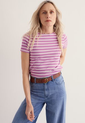 Kobieta o długich blond włosach, ubrana w fioletowo-białą koszulkę w paski, włożoną do wysokich niebieskich dżinsów z brązowym paskiem.