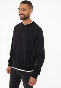 Czarny sweter z okrągłym dekoltem, z długimi rękawami, ściągaczami przy mankietach i u dołu. Noszony na białej koszulce z gładkiej bawełnianej mieszanki.