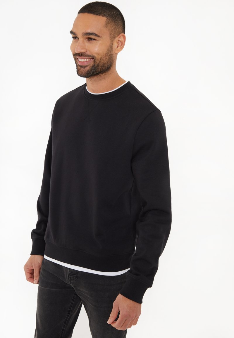 Czarny sweter z okrągłym dekoltem, z długimi rękawami, ściągaczami przy mankietach i u dołu. Noszony na białej koszulce z gładkiej bawełnianej mieszanki.