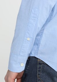 Světle modrá košile s dlouhým rukávem a bílými knoflíky, nošená s tmavě modrými denimovými džíny, ukazující detaily švů a lemu.