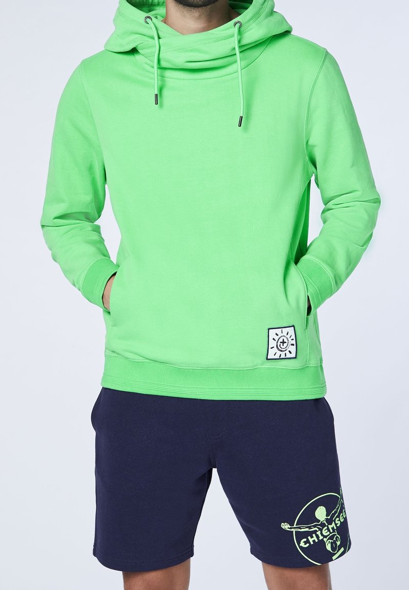 Personne portant un sweat à capuche vert vif avec des poches avant et un short bleu marine avec un logo jaune néon sur la jambe gauche.
