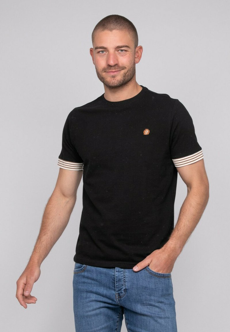 T-shirt noir à manches courtes avec des poignets rayés crème, col rond, et un petit logo brodé sur la poitrine. Le tissu semble lisse et ajusté.