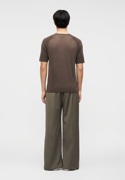 Άτομο που φαίνεται από πίσω, φοράει καφέ κοντομάνικο πουκάμισο και φαρδιά γκριζοκαφέ παντελόνια, με φαρδιά μπατζάκια, μπροστά σε απλό φόντο.