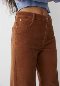 Bruna kordtbyxor med vertikalt ribbad textur, med två framfickor och metallknappsdetaljer. Hög midja.