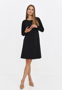 Czarna sukienka z długim rękawem i okrągłym dekoltem, dopasowaną górą, rozkloszowanym dołem oraz guzikami na ramionach. Gładki materiał, długość do kolan.