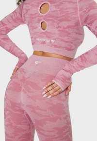 Femme portant un crop top à manches longues motif camouflage rose et un legging taille haute avec "Squatproof" et un logo, mains sur les hanches.