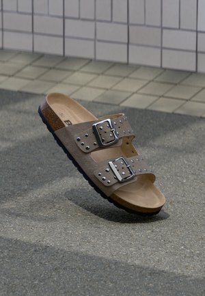 Καφέ δερμάτινα σανδάλια τύπου slide με δύο ρυθμιζόμενες μεταλλικές πόρπες και διακοσμητικά με καρφιά, με πάτο φτιαγμένο από φελλό και μαύρη λαστιχένια σόλα.