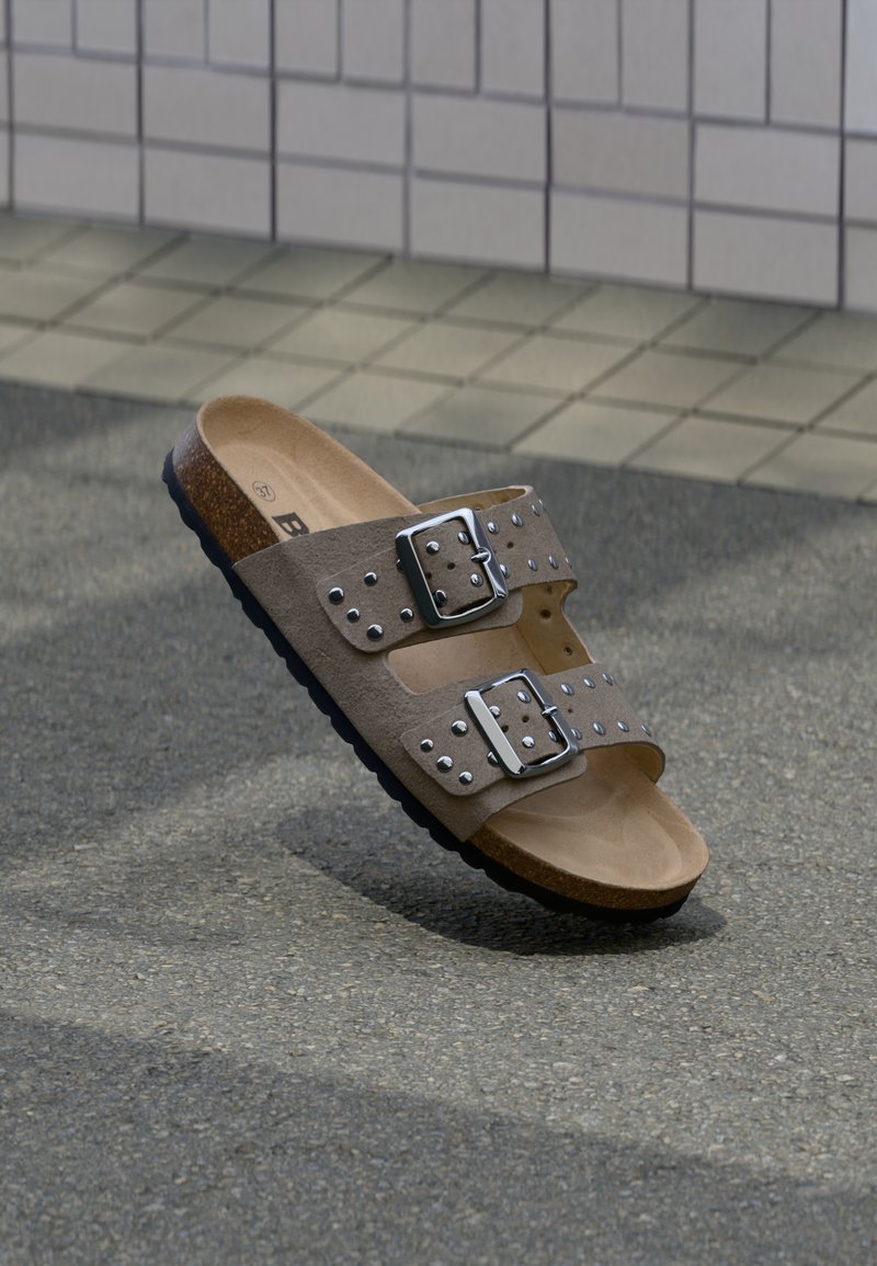 Καφέ δερμάτινα σανδάλια τύπου slide με δύο ρυθμιζόμενες μεταλλικές πόρπες και διακοσμητικά με καρφιά, με πάτο φτιαγμένο από φελλό και μαύρη λαστιχένια σόλα.