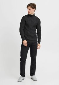 Schwarzer, gestrickter Rollkragenpullover mit strukturiertem Muster, kombiniert mit schwarzen Jeans und hellgrauen Sneakers, auf einem schlichten weißen Hintergrund.