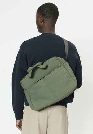 Vyras, dėvintis tamsiai mėlyną megztinį ir smėlio spalvos kelnes, per petį neša žalią rankinę baltame fone.