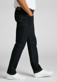 Pantalones vaqueros de mezclilla azul oscuro con corte ajustado, con costuras en contraste, cinco bolsillos y una etiqueta de cuero marrón en la cintura.