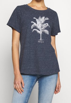 Personne portant un t-shirt bleu marine à manches courtes avec un motif de palmier blanc et le texte "ROXY", assorti à un jean bleu clair.