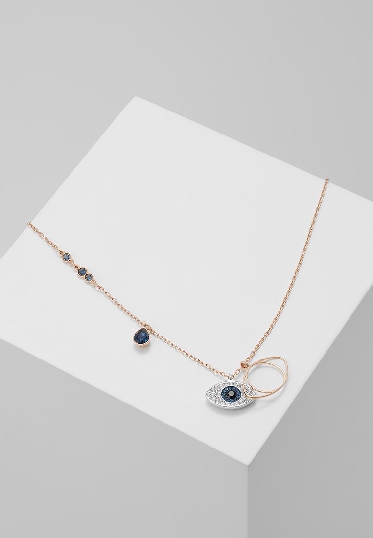 Collier délicat en or rose avec pendentif œil protecteur en pierres précieuses bleues et blanches et petites pierres bleues le long de la chaîne, présenté sur un bloc blanc.