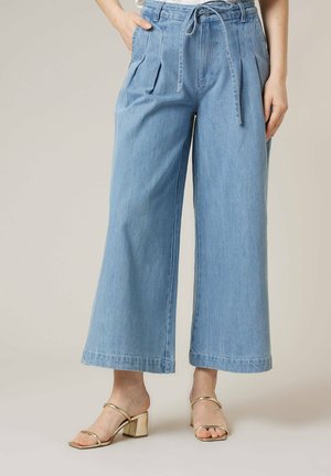 Vrouw draagt lichtblauwe, wijde, cropped spijkerbroeken met een trekkoord in de taille en gouden sandalen met blokhak en bandjes.