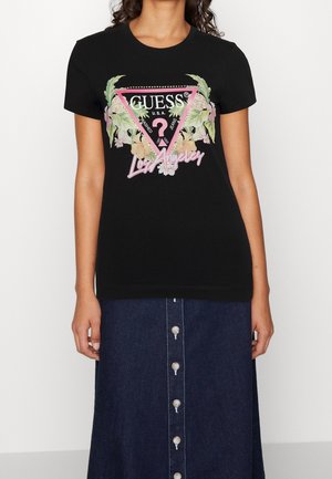 Kobieta w czarnym T-shircie Guess z różowym kwiatowym logo i napisem „Los Angeles”, połączonym z ciemną, zapinaną na guziki spódnicą dżinsową.
