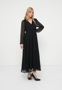 Frau steht mit einer Hand auf der Hüfte, trägt ein langes schwarzes Kleid mit transparenten Ärmeln und schwarze High Heels vor einem schlichten weißen Hintergrund.