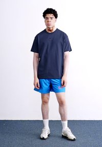 Tamsiai mėlynas Nike marškinėliai, mėlynos sportinės šortai ir balti kojinės. Avalynė turi šviesiai pilką ir baltą dizainą su tekstūriniais akcentais.