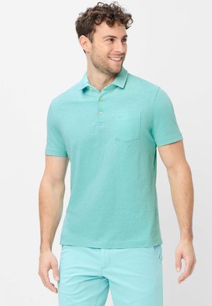 Lachende man met een licht turquoise poloshirt met borstzak en bijpassende broek, staand tegen een witte achtergrond.
