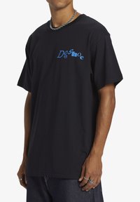 Man in een zwart oversized T-shirt met een blauw 'DC shoe' logo op de borst, gecombineerd met een donkere spijkerbroek en zilveren ringen en een ketting.