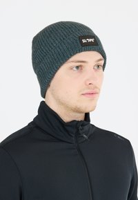 Mörkgrön ribbad mössa med en svart "SLOPE"-logotyppatch, bärs med en svart jacka med hög krage. Texturen verkar vara stickad.