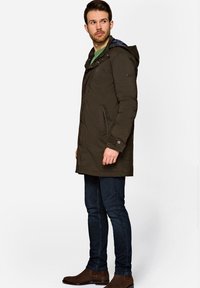 Oliwkowy park z kapturem, zapinany na zamek i guziki, z bocznymi kieszeniami i pikowaną teksturą, noszony na niebieskie dżinsy i brązowe buty.